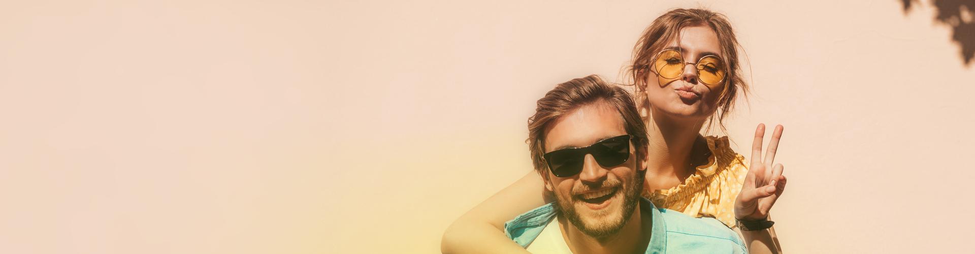 Ein junger Mann mit Sonnenbrille lächelt fröhlich in die Kamera und trägt eine Frau Huckepack Ein junger Mann mit Sonnenbrille lächelt fröhlich in die Kamera und trägt eine Frau Huckepack