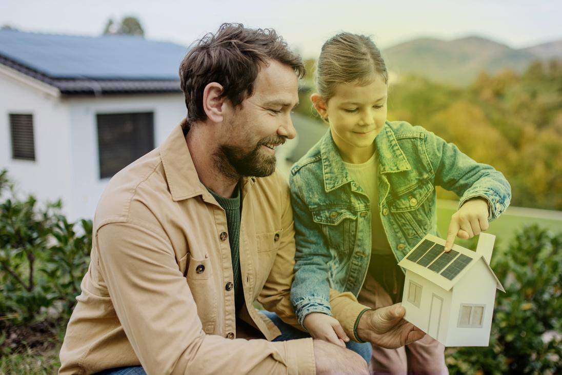 Ein Mann und ein kleines Mädchen stehen im Garten und schauen sich ein Modellhaus mit Photovoltaik auf dem Dach an. Ein Mann und ein kleines Mädchen stehen im Garten und schauen sich ein Modellhaus mit Photovoltaik auf dem Dach an.