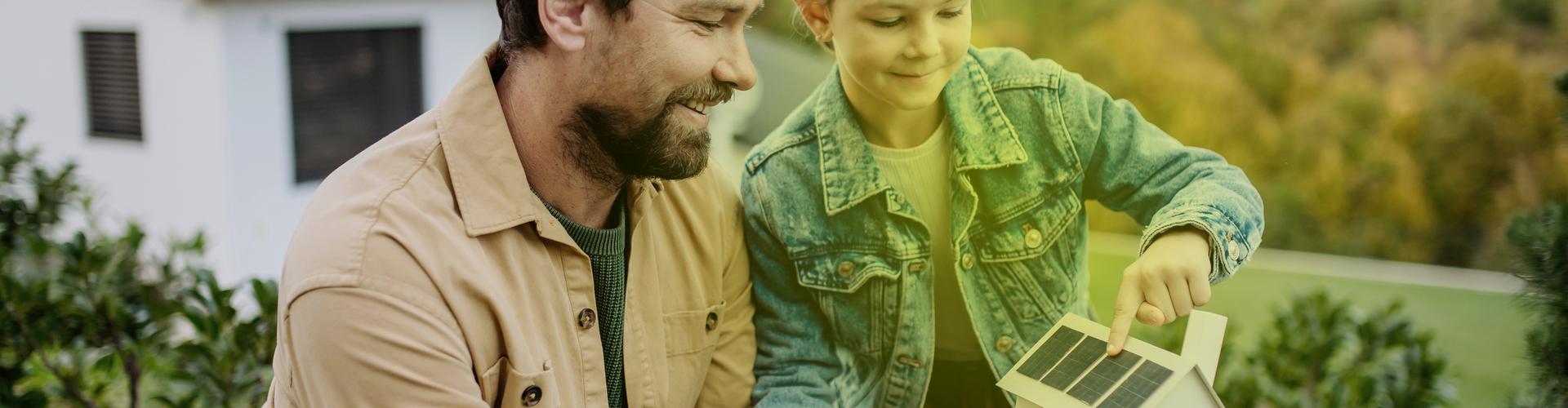 Ein Mann und ein kleines Mädchen stehen im Garten und schauen sich ein Modellhaus mit Photovoltaik auf dem Dach an. Ein Mann und ein kleines Mädchen stehen im Garten und schauen sich ein Modellhaus mit Photovoltaik auf dem Dach an.