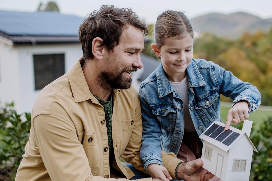 Ein Mann und ein kleines Mädchen stehen im Garten und schauen sich ein Modellhaus mit Photovoltaik auf dem Dach an. Ein Mann und ein kleines Mädchen stehen im Garten und schauen sich ein Modellhaus mit Photovoltaik auf dem Dach an.