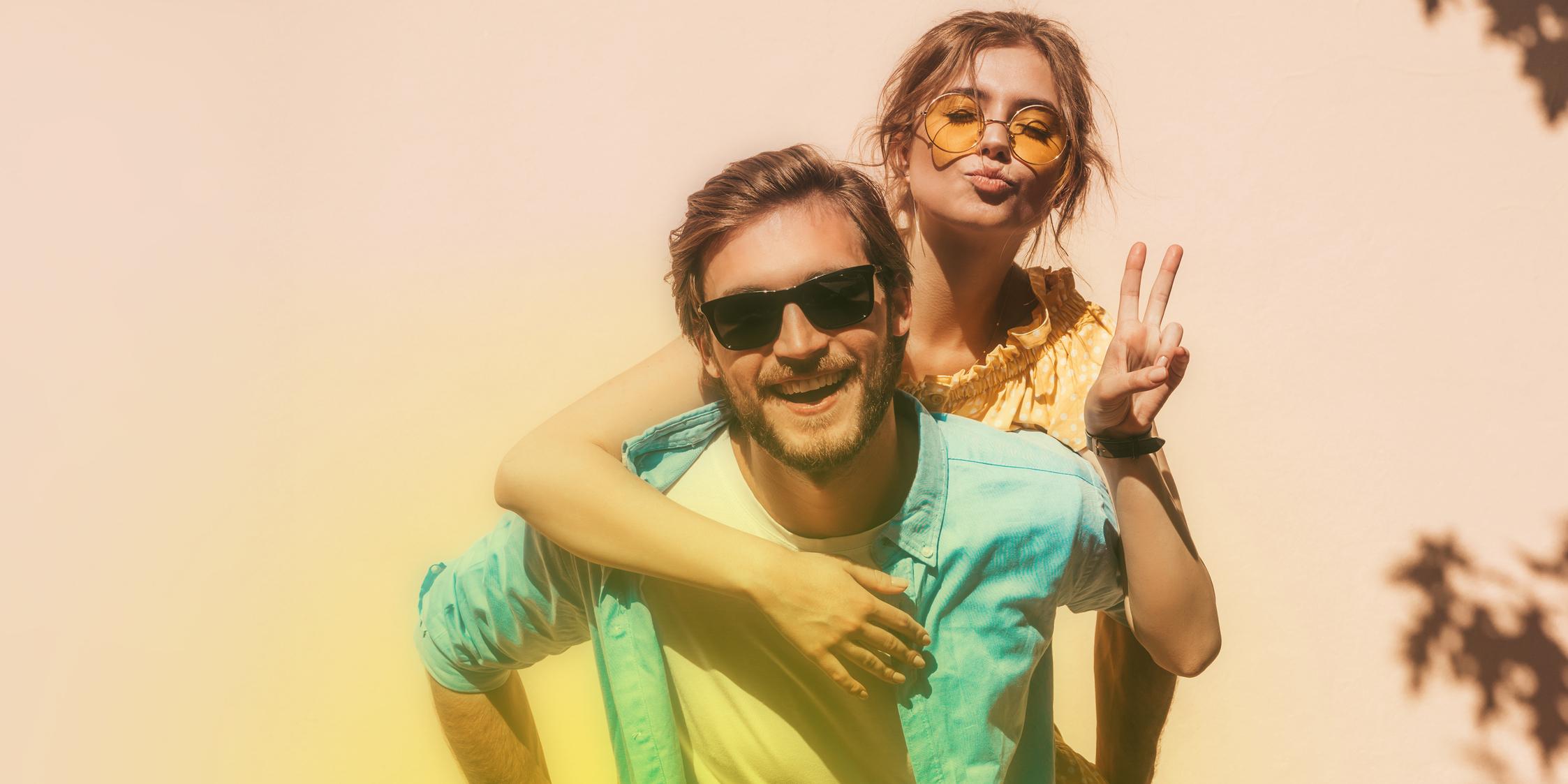 Ein junger Mann mit Sonnenbrille lächelt fröhlich in die Kamera und trägt eine Frau Huckepack Ein junger Mann mit Sonnenbrille lächelt fröhlich in die Kamera und trägt eine Frau Huckepack